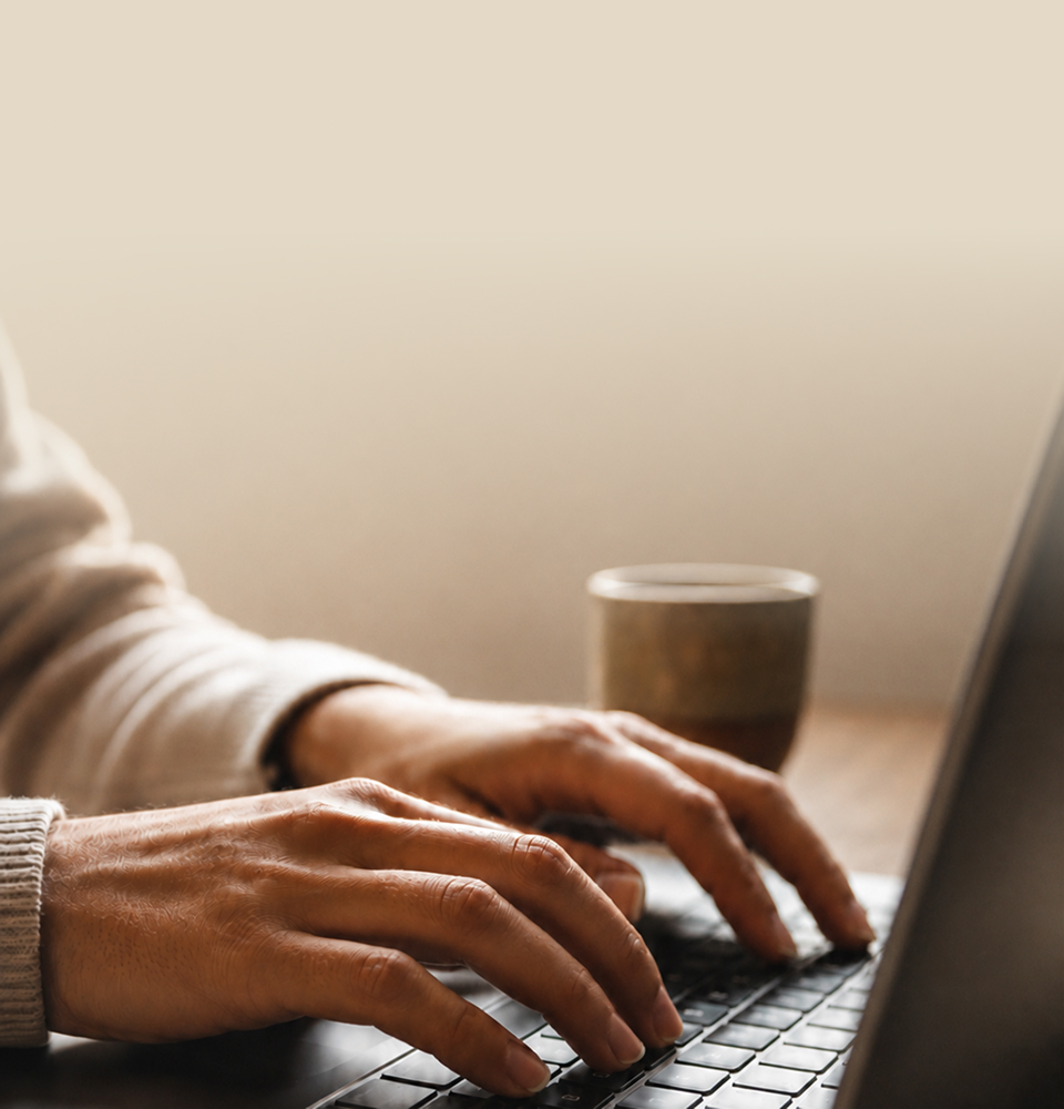 Hands typing on a laptop beside a coffee cup in warm window light